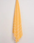 A yellow Turkish towel with white woven diagonal stripes and fringed edges, hanging from a hook against a plain off-white wall.