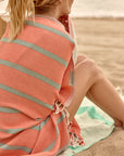 Lane Turkish towel in Peach Cream – seated with back turned, wrapped in towel on another Turkish towel by the shore