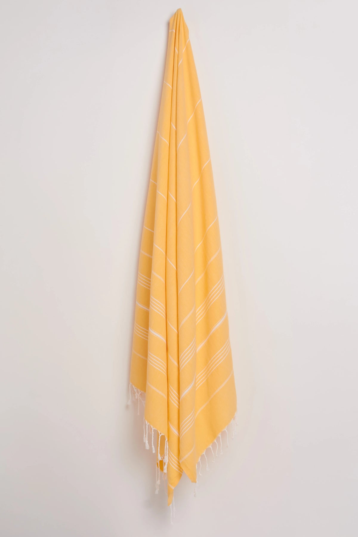 A yellow Turkish towel with white woven diagonal stripes and fringed edges, hanging from a hook against a plain off-white wall.