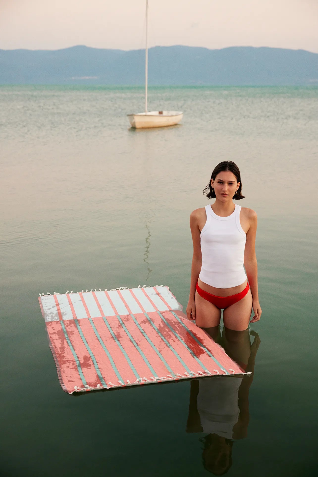 Lane Turkish towel in Peach Cream – floating on lake surface next to model with sailboat in background, towel absorbing water